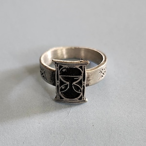 May include: A silver ring with an hourglass design. The hourglass is black and has a detailed design.