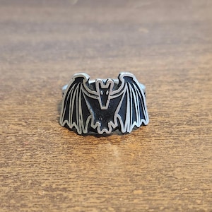 May include: A silver-toned metal ring featuring a flat, raised bat emblem. The bat design is detailed with black recessed areas that highlight the wings and body, creating a high-contrast look against the polished silver-colored metal edges.
