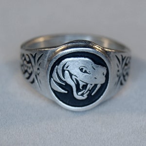 May include: A silver ring with a black enamel snake head design. The snake has a menacing expression with fangs showing. The ring band is decorated with a floral pattern.
