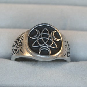Puede incluir: Un anillo de plata con un diseño de esmalte negro de un símbolo de triple luna y un nudo. El anillo tiene un diseño detallado en la banda.