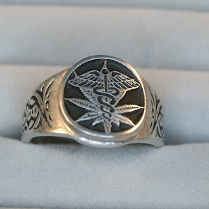 May include: A silver ring with a black and white design of a snake wrapped around a marijuana leaf. The ring is set in a round bezel.