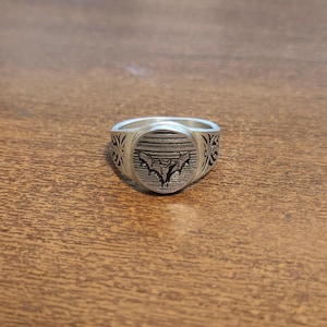 Może przedstawiać: Srebrny sygnet z szczegółowym emblematem nietoperza na okrągłej tarczy. Pierścień ma grawerowany wzór na obrączce i polerowane wykończenie. Pierścień jest prezentowany na drewnianej powierzchni.