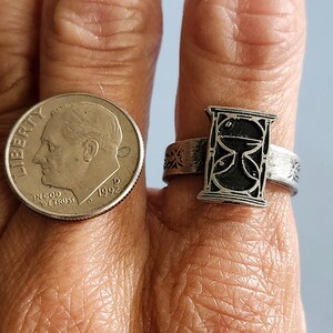 May include: A silver ring with an hourglass design. The hourglass is black and white and is set in a silver band. The band is decorated with a floral design.