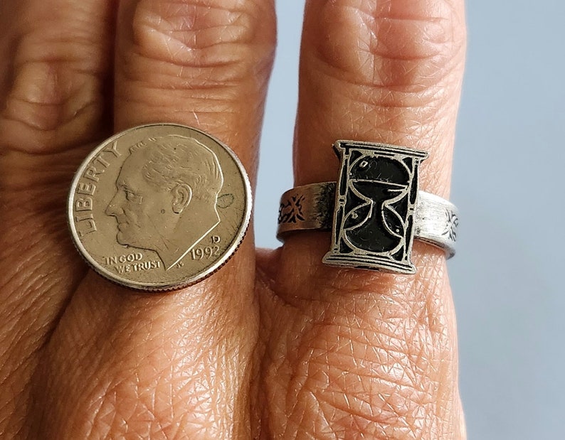 May include: A silver ring with an hourglass design. The hourglass is black and white and is set in a silver band. The band is decorated with a floral design.