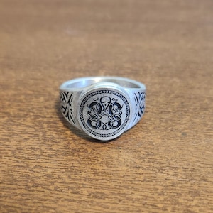 May include: Silver ring with a circular face featuring a black, stylized design. The ring's band has intricate patterns on the sides. The ring is set against a wooden surface.