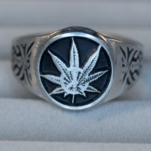 May include: A silver ring featuring a black and white marijuana leaf design. The ring band has a detailed design.