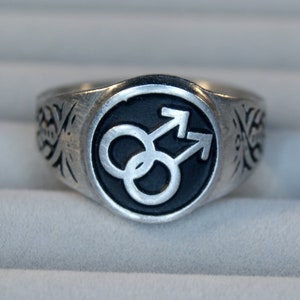 May include: A silver ring with a black enamel inlay featuring a stylized symbol of two interlocking arrows.