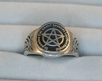 May include: A silver ring with a black pentagram and the inscription "Domine Del Nostri Satanas Lucifer Excelsis" around the pentagram.