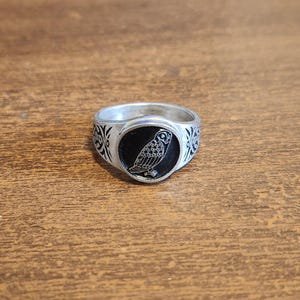 May include: Silver-toned ring with a black oval center featuring a detailed owl illustration. The ring has decorative patterns on the sides and is set against a wooden surface. A unique piece of jewelry.