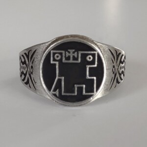 May include: A silver ring with a black enamel design of a stylized figure with a cross on top. The ring has a decorative band with a floral pattern.