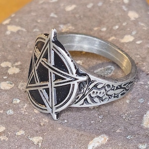 Puede incluir: Un anillo plateado con un diseño de pentagrama negro. El anillo tiene un patrón floral detallado en la banda. El anillo está sobre una superficie de piedra gris.