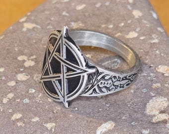 Puede incluir: Un anillo plateado con un diseño de pentagrama negro. El anillo tiene un patrón floral detallado en la banda. El anillo está sobre una superficie de piedra gris.