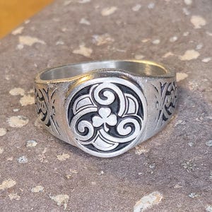 May include: A silver-colored ring with an engraved Celtic knot design. The ring features a central circular emblem with a three-leaf clover and swirling patterns. The band has additional decorative elements.