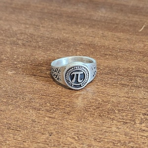 May include: Silver ring featuring the mathematical symbol Pi (π) in a black circle, surrounded by numbers and decorative patterns. The ring is on a wooden surface.