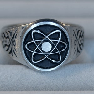May include: A silver ring with a black atom symbol on the top. The ring has a detailed design on the band.