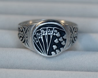 Puede incluir: Un anillo de plata con un diseño de esmalte negro de cartas de juego, incluyendo el As, el Rey, la Reina, el Jack y el 10 de tréboles.