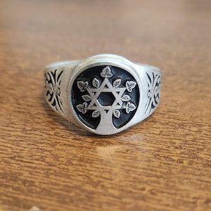 May include: A silver ring with a black circular center featuring a Star of David tree design. The ring's band has decorative floral patterns on each side. The ring is displayed on a wooden surface.