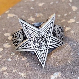 Puede incluir: Anillo plateado con un diseño de pentagrama en relieve. El pentagrama presenta una cabeza de lobo estilizada en el centro. La banda del anillo está decorada con un patrón ornamentado. El anillo está sobre una superficie de piedra gris texturizada.