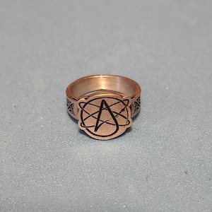Könnte beinhalten: Ein silberner Ring mit einem schwarzen Symbol eines 'A' in einem Kreis. Der Ring ist mit einem Muster um den Band graviert.
