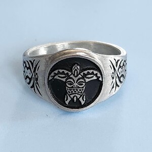 May include: Silver-toned ring featuring a black, circular centerpiece with a tribal design. The ring's band has etched patterns on the sides. The ring is set against a light blue background.