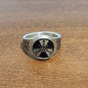 May include: Silver-toned ring featuring a black circular emblem with a skull and crossbones design, surrounded by a radiation symbol. The ring has an engraved pattern on the band. The ring is displayed on a wooden surface.