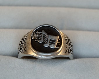 May include: A silver ring with a black circular inlay featuring a silver musical note design.