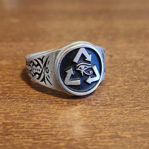 Puede incluir: Anillo plateado con una cara circular. La cara tiene un fondo negro con un símbolo de reciclaje blanco y un ojo en el centro. La banda del anillo tiene un diseño tribal grabado. El anillo está sobre una superficie de madera.