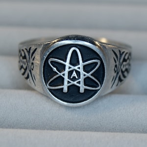 May include: A silver ring featuring a black enamel atom symbol with the letter 'A' inside. The ring has a decorative design on the sides.