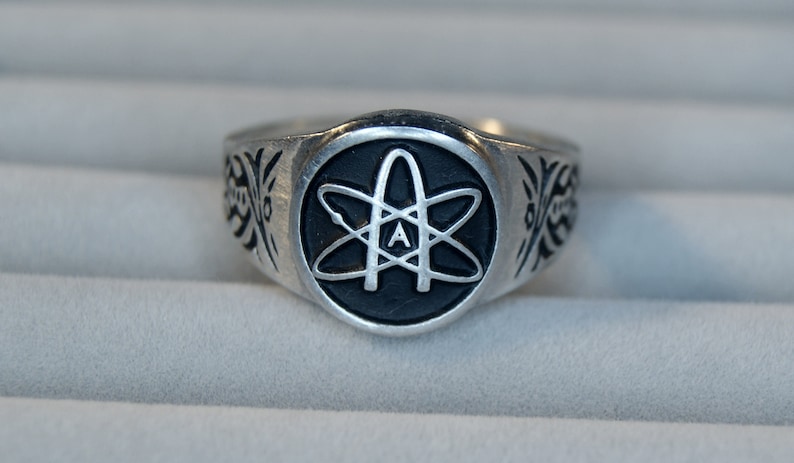 May include: A silver ring featuring a black enamel atom symbol with the letter 'A' inside. The ring has a decorative design on the sides.