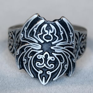 Puede incluir: Un anillo de plata con un diseño detallado de una araña en el centro. La araña está rodeada de intrincados patrones florales.