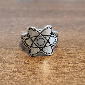 May include: Silver-toned ring featuring an atom design. The ring's face showcases a central circle with intersecting lines, surrounded by a decorative band. The ring is made of a metallic material and sits on a wooden surface.