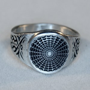 May include: A silver ring with a black spiderweb design on the top. The ring is engraved with a tribal design on the sides.