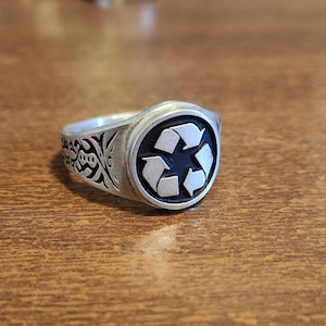 Puede incluir: Un anillo plateado con una cara circular con un símbolo de reciclaje negro. El anillo tiene un diseño ornamentado en la banda. El anillo está sobre una superficie de madera.