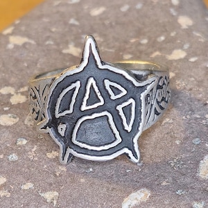 Puede incluir: Anillo plateado con un prominente símbolo de anarquía en un diseño audaz y estilizado. El anillo tiene una banda detallada y texturizada con patrones florales y geométricos. El anillo está sobre un fondo neutro y texturizado.