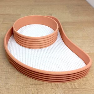 Puede incluir: Una bandeja escalonada de color melocotón con una superficie texturizada blanca. La bandeja tiene una sección superior circular y una base curva y alargada. El diseño es moderno y minimalista.