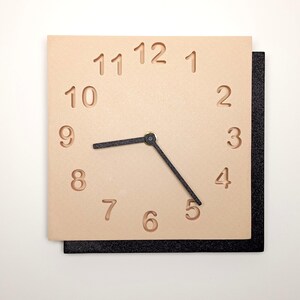 May include: Square wall clock with a light tan face and black hands. The numbers are cut out of the face. The clock is mounted on a black square base. The clock hands point to 8:40.