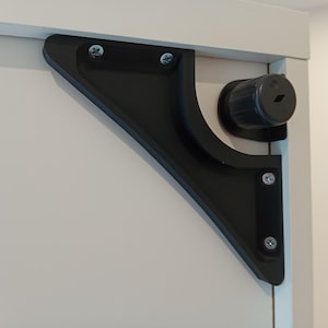 Op de afbeelding: Een zwarte hoekbeugel met een gebogen ontwerp, gemonteerd op een wit oppervlak. De beugel is bevestigd met zilveren schroeven en heeft een cilindrisch onderdeel. Het ontwerp is modern en functioneel.