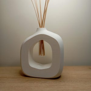Può includere: Un diffusore di oli essenziali bianco, in stile moderno, con un design aperto unico. Il diffusore contiene diversi bastoncini di legno e si trova su una superficie di legno. Un oggetto decorativo per la casa.