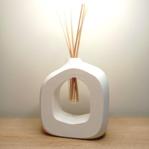 Pode incluir: Um difusor de óleos essenciais branco, de estilo moderno, com um design aberto único. O difusor contém várias varetas de madeira e está colocado sobre uma superfície de madeira. É um artigo decorativo para a casa.