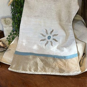 Op de afbeelding: Een witte linnen theedoek met een geborduurd zonontwerp in blauw en bruin. De handdoek heeft een blauwe en bruine rand en een witte gehaakte rand.