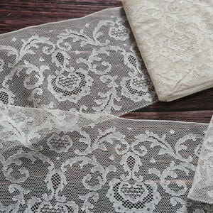 Puede incluir: Tela de encaje marfil con un patrón floral. El encaje está dispuesto en tiras y piezas dobladas, mostrando el intrincado diseño. La tela se coloca sobre un fondo de madera oscura, resaltando la delicada textura y el color.