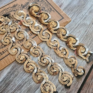 Pode incluir: Acabamento de lantejoulas douradas com um design em espiral e ornamentado. O acabamento é disposto sobre um fundo de papel vintage com texto francês e uma superfície de madeira. As lantejoulas são de cor dourada quente e o acabamento é provavelmente usado para embelezar.