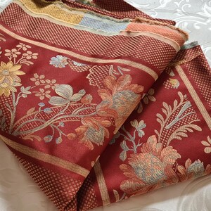 May include: Red fabric with a floral pattern in shades of gold, blue, and cream. The fabric has a subtle textured pattern and a striped border.