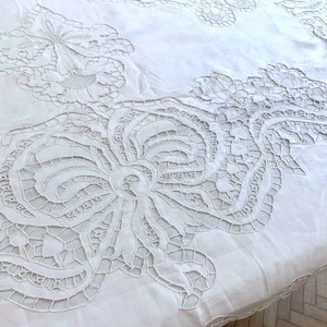 Puede incluir: Mantel de mesa de algodón blanco con intrincados diseños florales bordados. El mantel presenta un gran motivo floral central rodeado de patrones florales más pequeños.