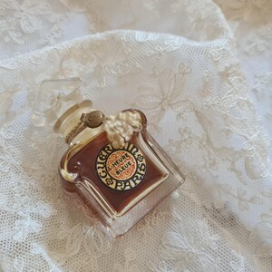 May include: A vintage glass bottle of perfume with a gold cap and a label that reads "L'Heure Bleue Paris". The bottle is sitting on a white lace fabric.