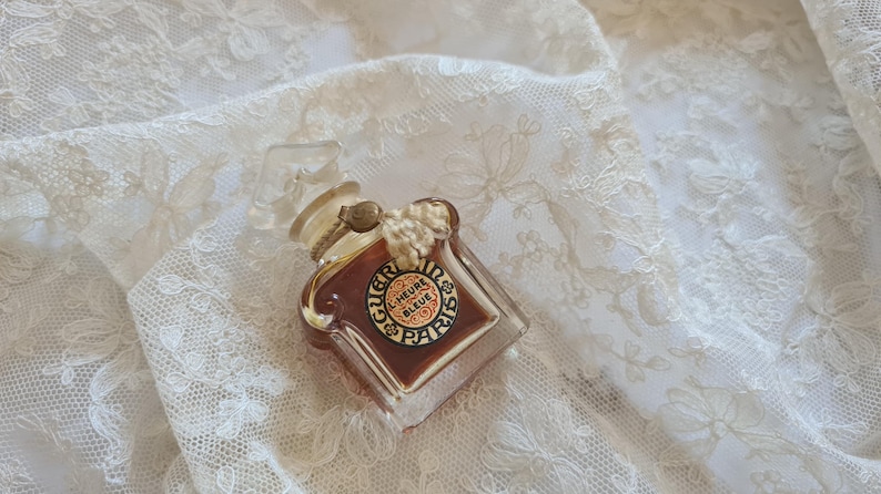 May include: A vintage glass bottle of perfume with a gold cap and a label that reads "L'Heure Bleue Paris". The bottle is sitting on a white lace fabric.