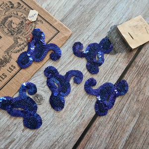 May include: Five navy blue sequin appliques with a swirling design are arranged on a wooden surface. A vintage chocolate box and a small tag are also present. The appliques are embellished with sequins and beads.