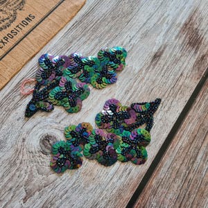 May include: Two clusters of iridescent sequin appliques in shades of green, purple, and black, arranged on a wooden surface. The appliques are embellished with small black beads. A vintage paper fragment is in the background.