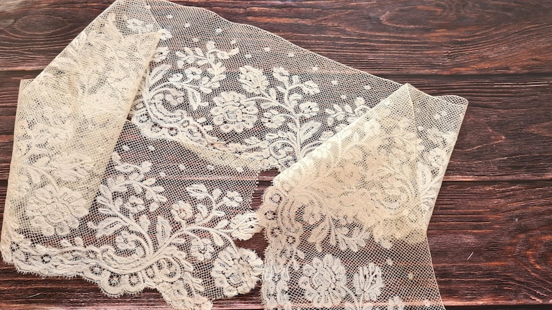 Puede incluir: Tela de encaje marfil con un dise&ntilde;o floral. El encaje est&aacute; doblado, revelando los intrincados patrones de flores y hojas. La tela est&aacute; colocada sobre una superficie de madera oscura.