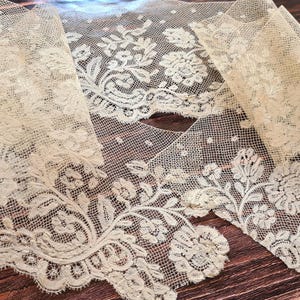 Puede incluir: Tela de encaje color marfil con dise&ntilde;os florales y de hojas. El encaje se muestra sobre una superficie de madera oscura, resaltando su delicado dise&ntilde;o y bordes festoneados. La tela est&aacute; doblada en secciones.
